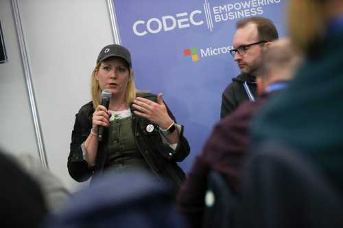 A woman speaking on a microphone to a crowd while a man looks on.